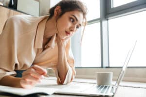 Clinician preparing for a board exam—focused on a laptop with pen, notebook, and coffee nearby in bright window light.